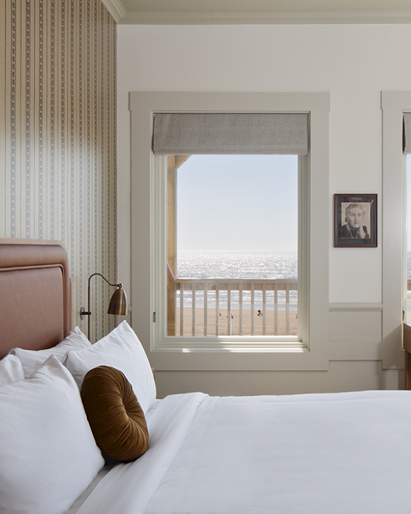A coastal-themed bedroom features a bed, pillow, framed photo, desk, and ocean view through windows, creating a serene atmosphere.
