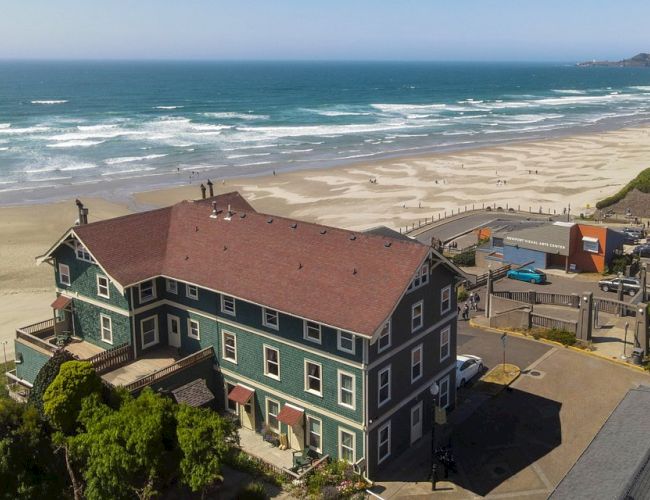 A large building with a red roof is situated near a beach, with waves and a clear sky in the background.