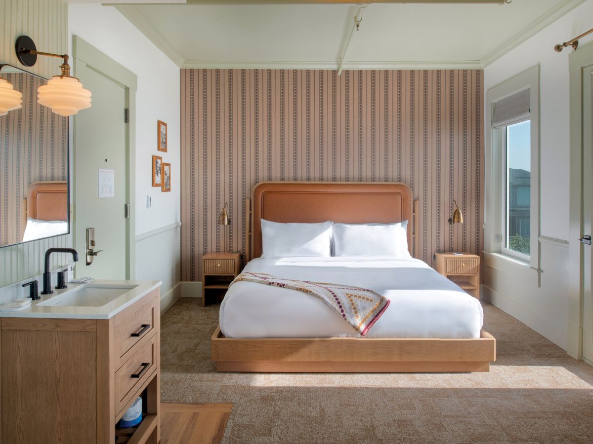 A cozy bedroom with a large bed, patterned wallpaper, and a bedside table. There's a small sink and a mirror on the left side.
