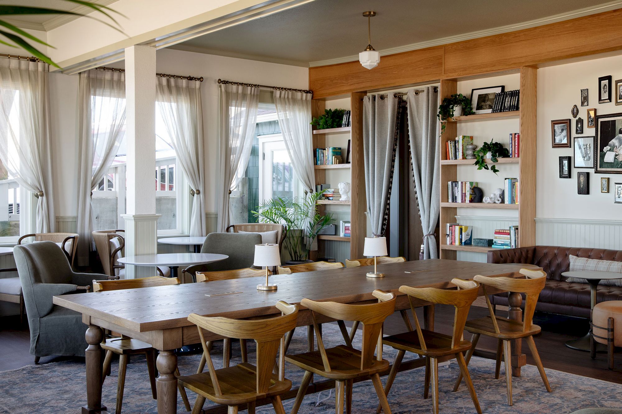 A cozy room featuring a long wooden table, chairs, couches, bookshelves, and framed photos on the wall, with natural light streaming in.