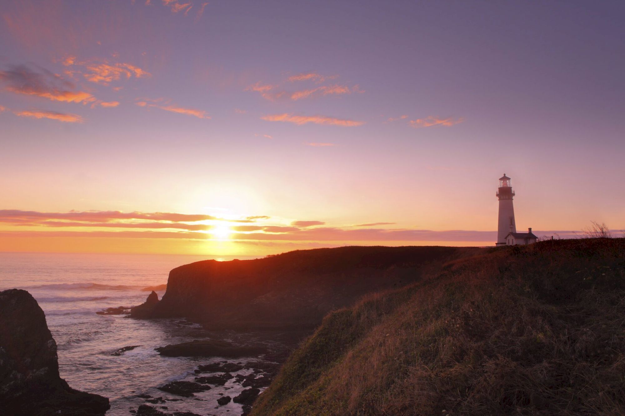 A lighthouse stands on a cliff overlooking the ocean at sunset, with a colorful sky and calm waters below.