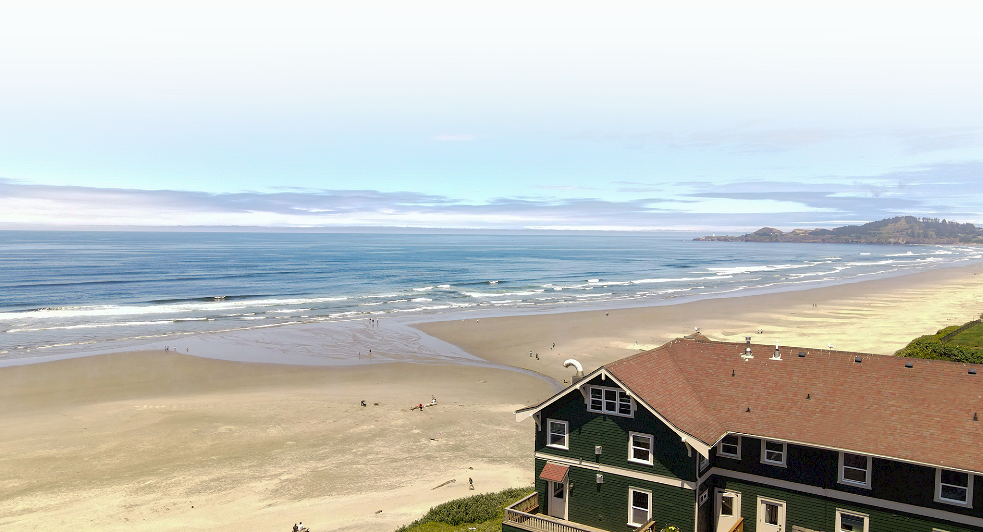 A coastal scene with a sandy beach, ocean waves, and a house with a red roof in the foreground under a partly cloudy sky.