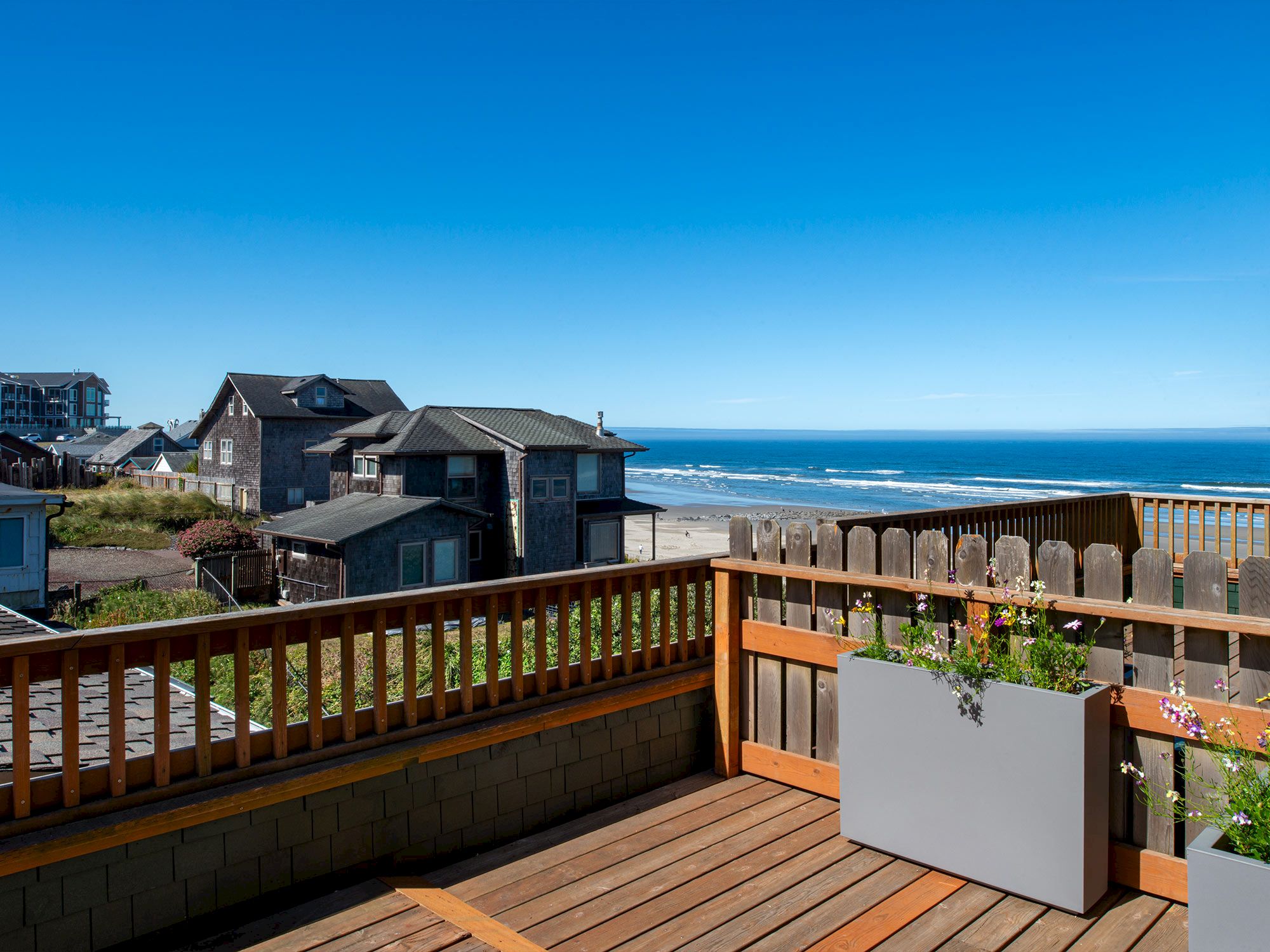 A seaside view from a wooden deck, with houses nearby and the ocean visible under a clear blue sky.