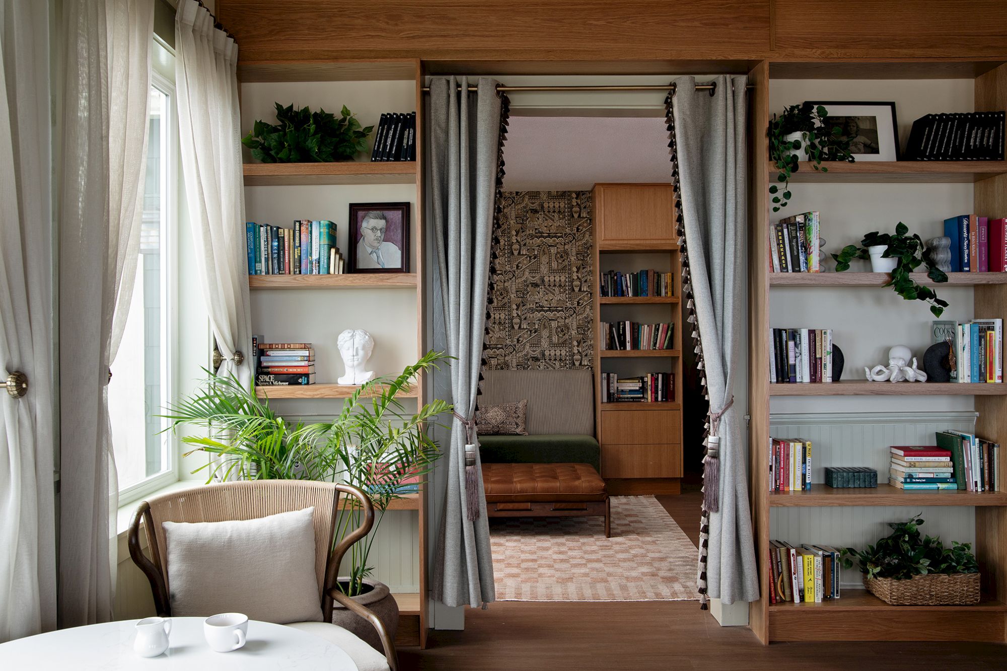 A cozy reading room with bookshelves, curtains, plants, a chair, and a glimpse into a second room with more shelves and seating.