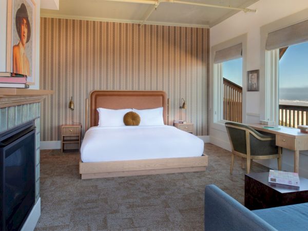 The image shows a modern bedroom with a bed, striped walls, a small desk, and a fireplace, with large windows offering a scenic view.