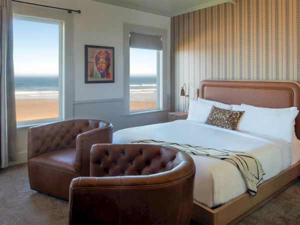 A cozy bedroom with a seaside view, featuring a large bed, two brown chairs, and a framed portrait on the wall.