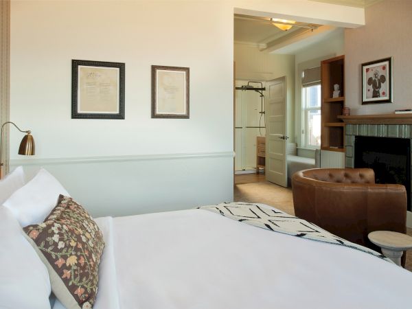 A cozy bedroom with a neatly made bed, artwork on the walls, a leather chair, a fireplace, and a doorway leading to another room.