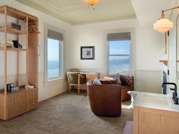 A cozy room with a leather chair, desk, bookshelf, and sink. Large windows offer an ocean view under soft, warm lighting.