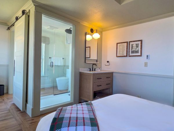 The image shows a hotel room with a bed, a glass shower, a small sink area, a chair, and framed pictures on the wall.