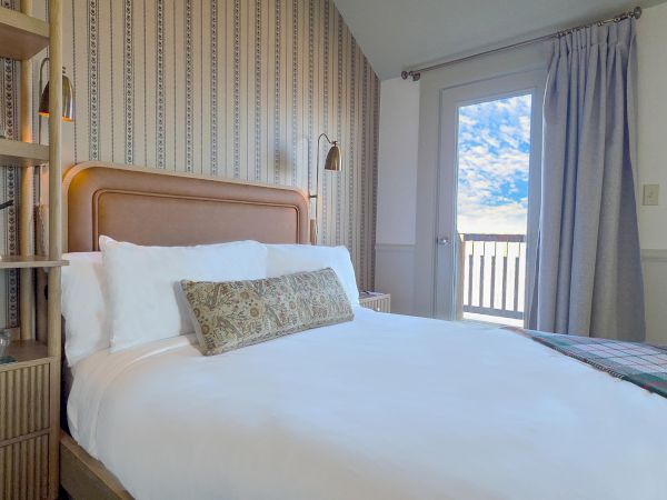 A cozy bedroom features a neatly made bed, a bookshelf, wall art, and a door with curtains leading to an outdoor view.