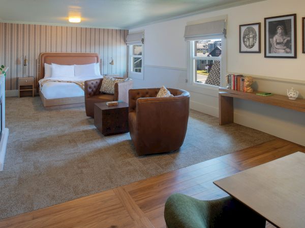 The image shows a spacious hotel room with a large bed, seating area, artwork, and a desk with books, blending comfort and style.