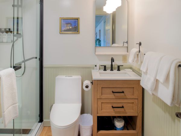 A small bathroom with a shower, toilet, sink, mirror, and towels. The decor includes a picture and light fixture above the sink area.
