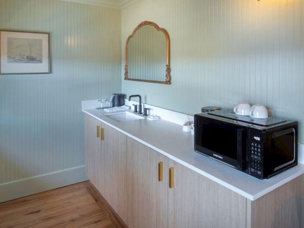 The image shows a small kitchenette with a microwave, cups, kettle, and mirror above a counter with cabinets in a light-colored room.