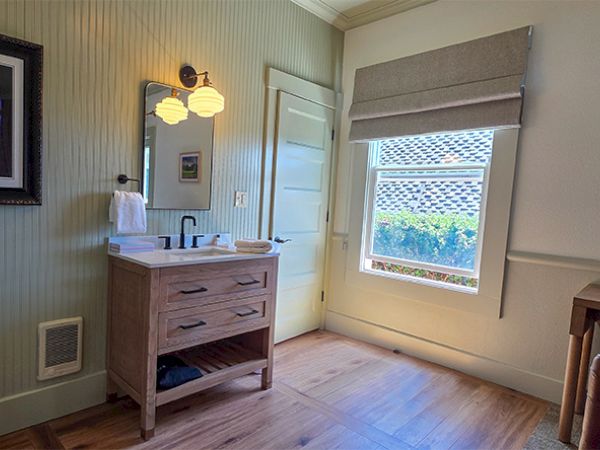 The image shows a bathroom with a wooden vanity, mirror, lighting, window with blinds, and wall art on a light-colored wall.