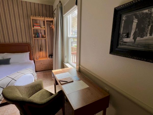The image shows a cozy room with a bed, a desk by the window, a chair, a bookshelf, and a framed picture on the wall.