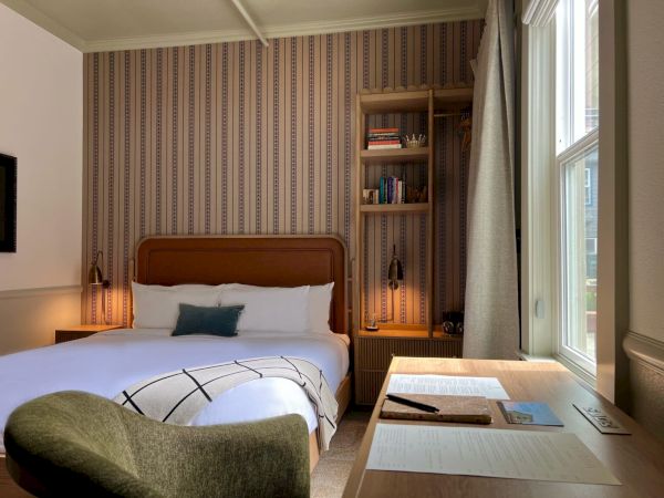 The image shows a cozy hotel room with a bed, armchair, desk, and window. The decor is warm with a striped wall, lamp lighting, and books.