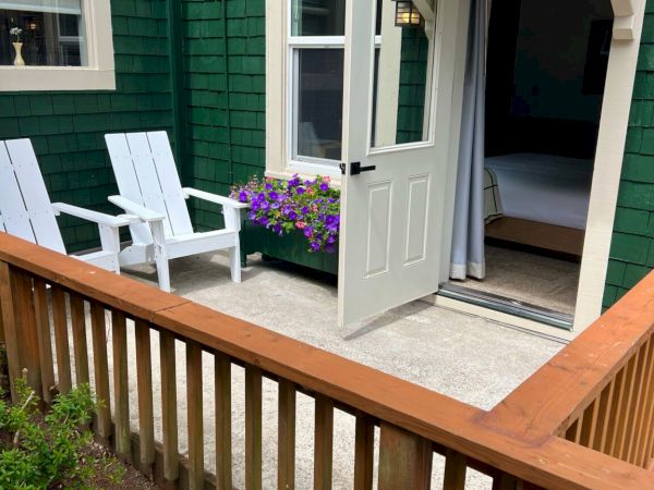A cozy patio with white chairs, a flower box, and a partially open door leading to a bedroom. Garden flowers bloom in the foreground.