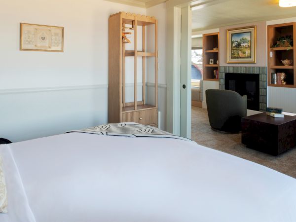 A cozy room with a bed, small table, armchair, wall art, shelving, and fireplace creates a comfortable, inviting atmosphere.