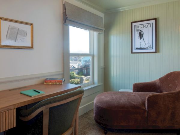 A cozy room with a desk, chair, window, framed pictures, and a chaise lounge under soft light.