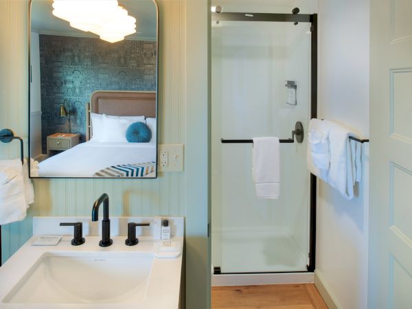 The image shows a bathroom with a sink, mirror, and a shower with glass doors. Towels hang nearby, and a glimpse of a bedroom is visible.