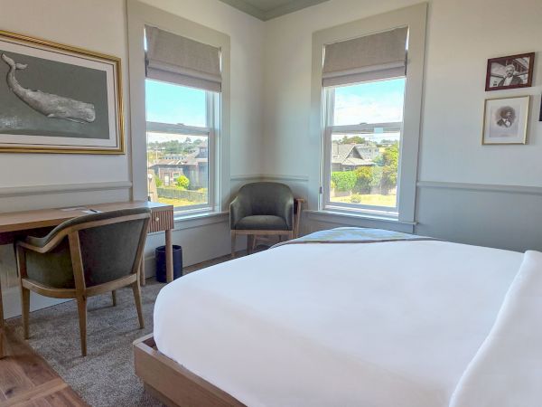 A cozy bedroom with a bed, desk, two chairs, and large windows showcasing a scenic view; artwork decorates the walls.