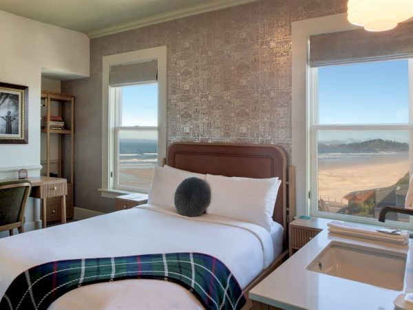 A cozy hotel room with a bed, plaid blanket, desk, and two windows showcasing a beach view and ocean in the distance.