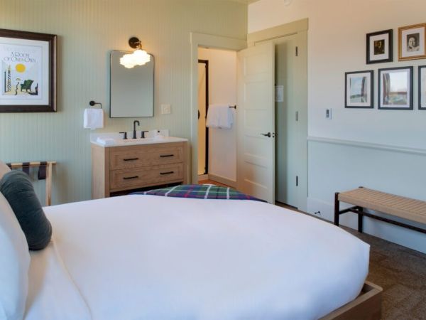 A cozy bedroom features a neatly made bed, a wooden bench, framed art on the walls, a sink with a mirror, and a door leading to a bathroom.