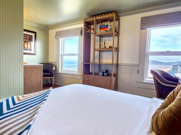 A cozy bedroom with ocean views, a comfy bed, patterned throw, and shelf with books. Two large windows let in natural light.