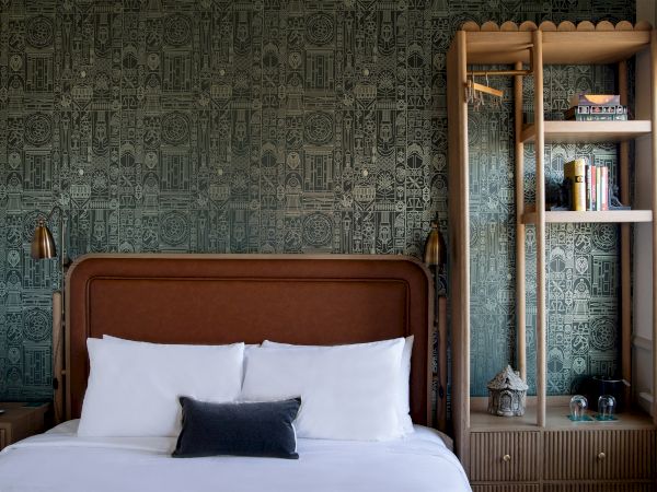 A cozy bedroom with a patterned wallpaper, bed with white pillows, eclectic lighting, and rustic shelving with books and decorative items.
