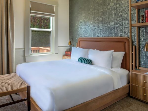 The image shows a cozy bedroom with a neatly made bed, decorative pillows, a window with blinds, and a bookshelf with books and decor.