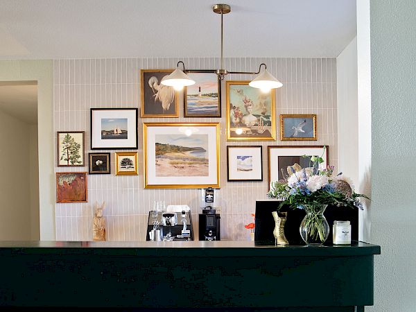 A dark green bar with a brass foot rail sits in front of a wall gallery of framed art; a couple of hanging lights illuminate the colorful collage above.