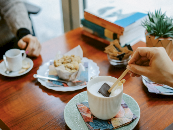 Two people are having coffee at a table with a muffin, books, a small plant, and a hand stirring a drink with a stick in a mug.