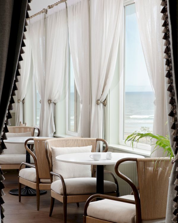 A cozy café interior with elegant chairs, round tables, and sheer white curtains framing large windows.