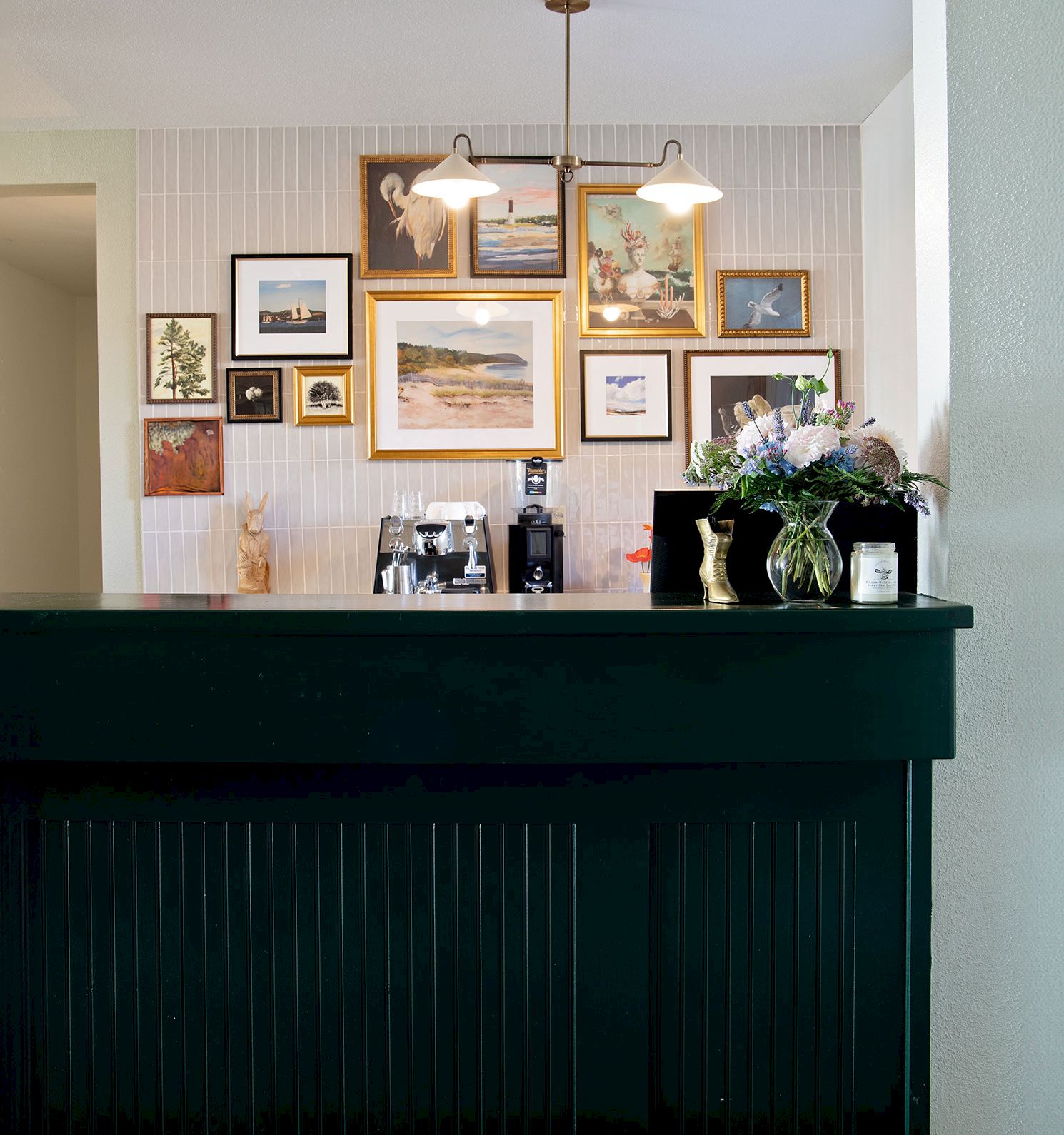A stylish home bar with a black counter, a gallery wall of framed art, pendant light, and a vase of flowers decorates the space.