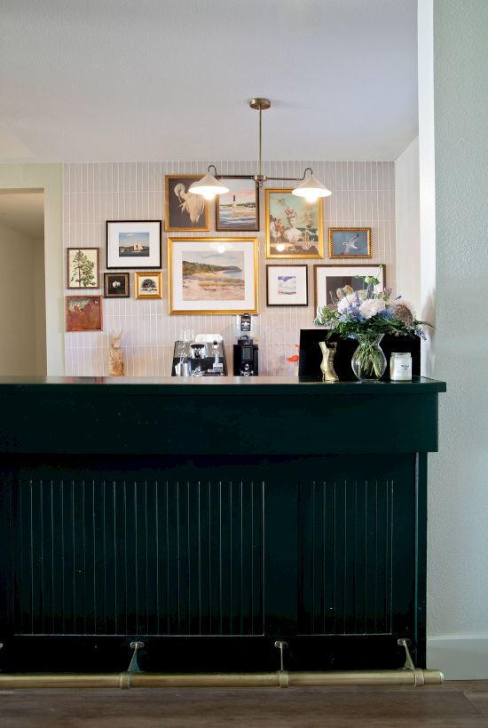 A stylish home bar with a black counter, a gallery wall of framed art, pendant light, and a vase of flowers decorates the space.