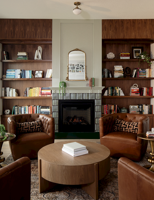 A cozy living room with leather chairs, a central coffee table, a fireplace, bookshelves, and decorative items.