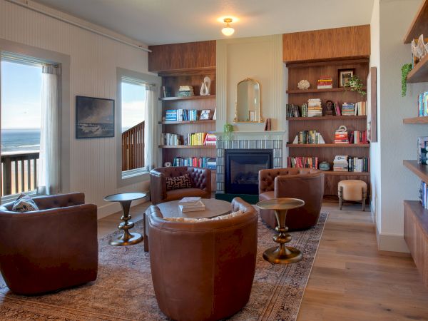 A cozy room with leather chairs, bookshelves, a fireplace, and ocean views through large windows, creating a relaxing atmosphere.