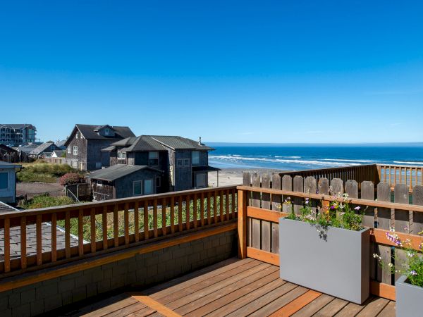Wooden deck with planter overlooks seaside homes, sandy beach, and blue ocean under a clear sky.