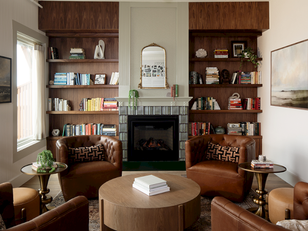 A cozy living room with a fireplace, bookshelves, leather chairs, and a round coffee table in a warmly lit, inviting setting.
