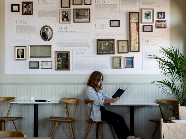 A person sits reading in a cafe with a wall display of framed photos and text about Sylvia Beach, surrounded by wooden chairs and tables.