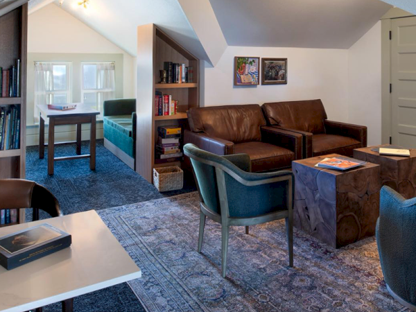 A cozy room features leather seating, shelves, tables, and a daybed. There's warm lighting, framed artwork, and a patterned rug on the floor.