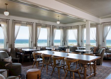 A cozy seaside restaurant interior with wooden tables, chairs, and large windows offering a view of the ocean.