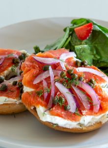 A plate with a bagel topped with cream cheese, smoked salmon, red onions, and capers, alongside a green salad with a tomato.
