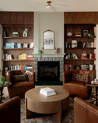 A cozy living room with a fireplace, surrounded by bookshelves, leather chairs, a round table, and artwork on the walls.
