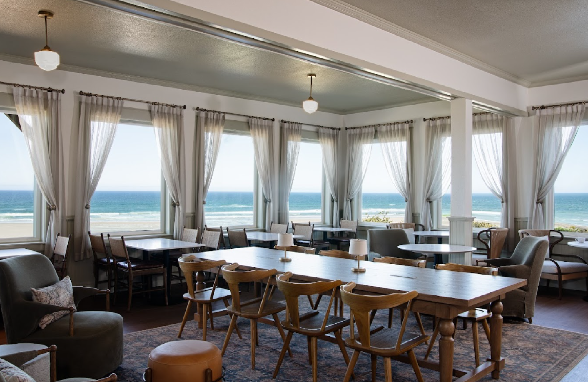 The image shows a cozy dining area with wooden tables, chairs, and large windows offering a view of the ocean in the background.