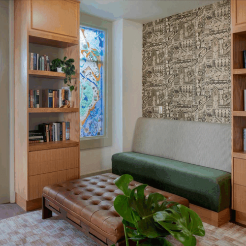 A cozy room with wooden shelves, books, a green bench, patterned wallpaper, a tufted ottoman, and a colorful stained glass window.