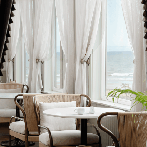 A cozy café with white curtains, a round table, and chairs overlooks the ocean through large windows, creating a serene atmosphere.