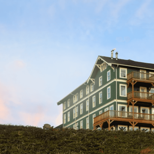 A large green house with white trim sits on a hill under a blue sky with clouds, featuring multiple balconies and wooden supports.