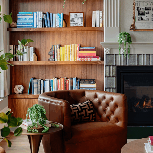 A cozy living room with a leather chair, bookshelves with colorful books, plants, and a fireplace, creating a warm atmosphere.