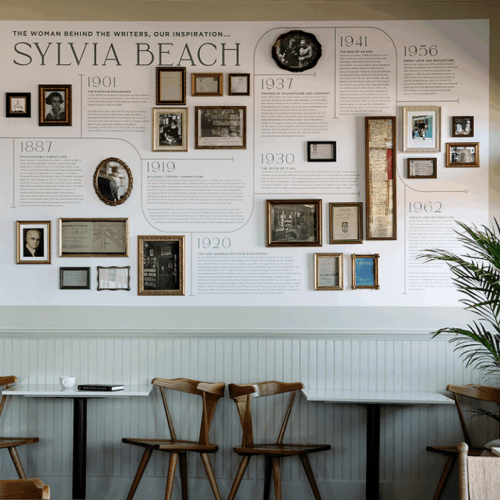 The image shows a wall display about Sylvia Beach, featuring her biography with framed photos and text in a cafe setting.